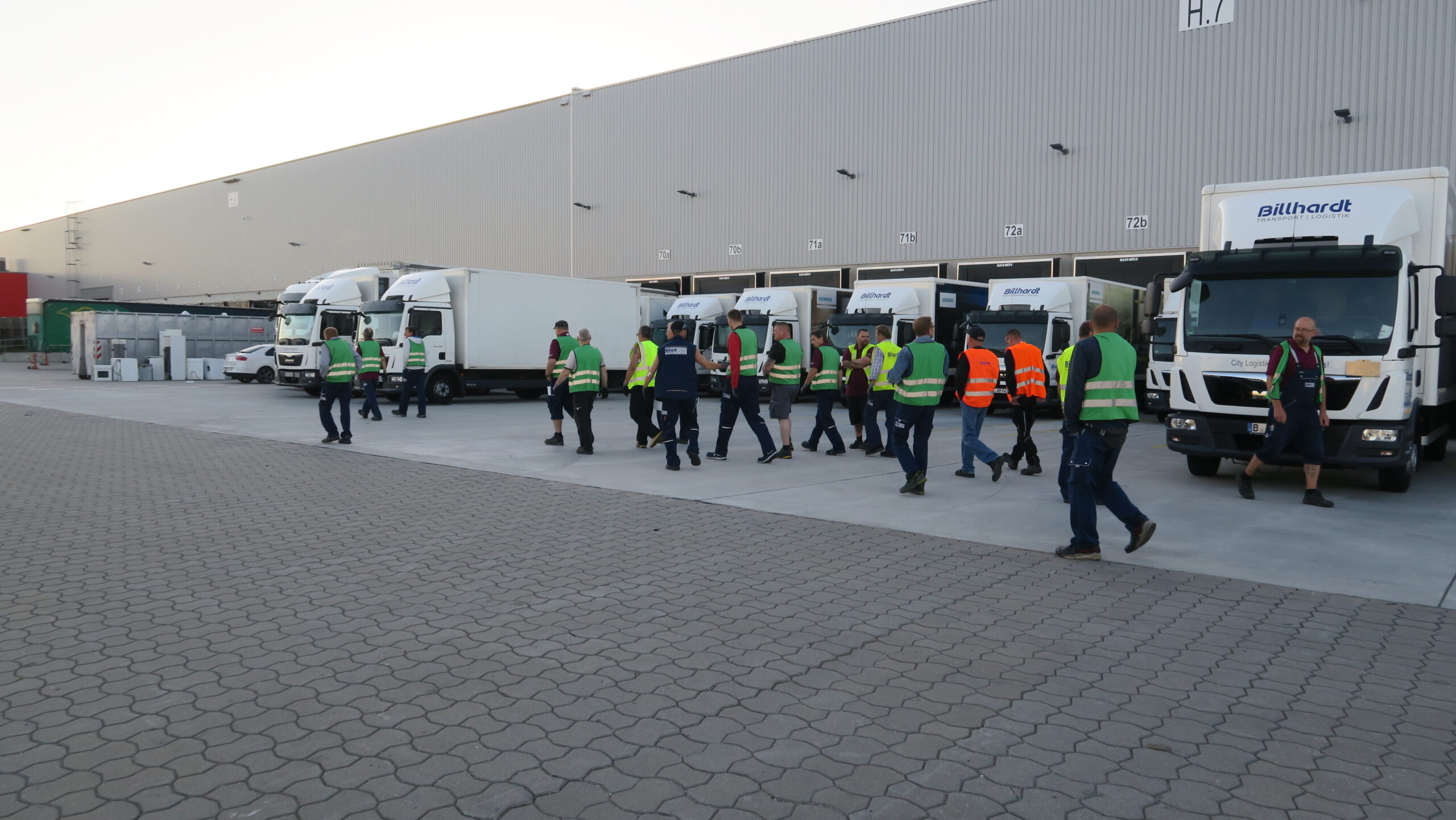 Gruppe von Arbeitern mit Schutzwesten und Helmen vor mehreren LKWs an einem Lagerhaus.
