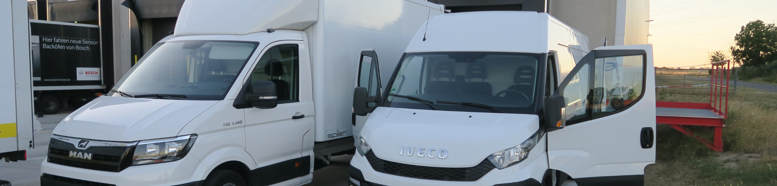 Zwei weiße Lieferwagen nebeneinander auf einem Feldweg bei Sonnenuntergang.