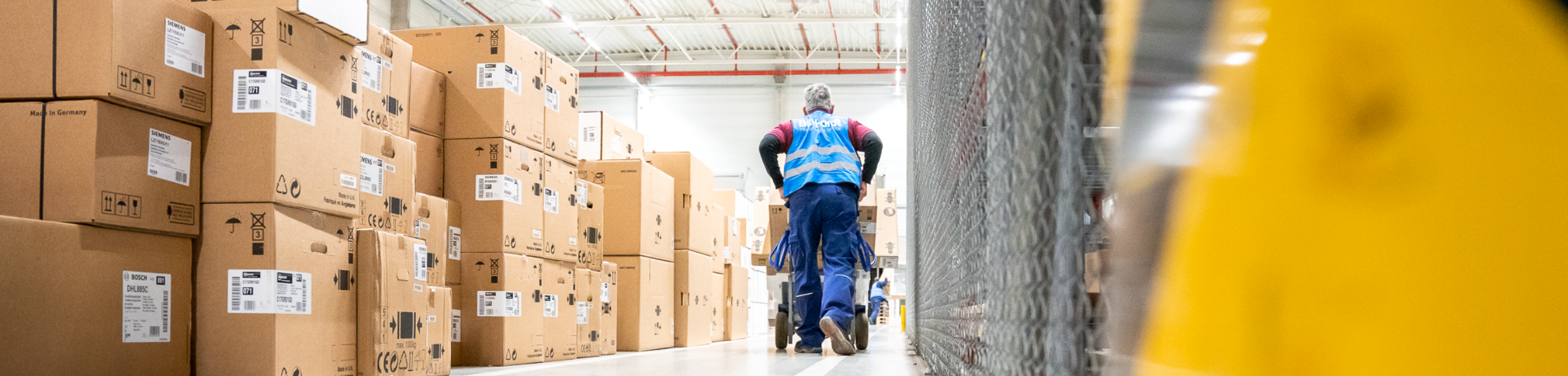 Lagerarbeiter mit Schutzhelm und Weste bewegt Stapel von Kartons in einem Lagerhaus.