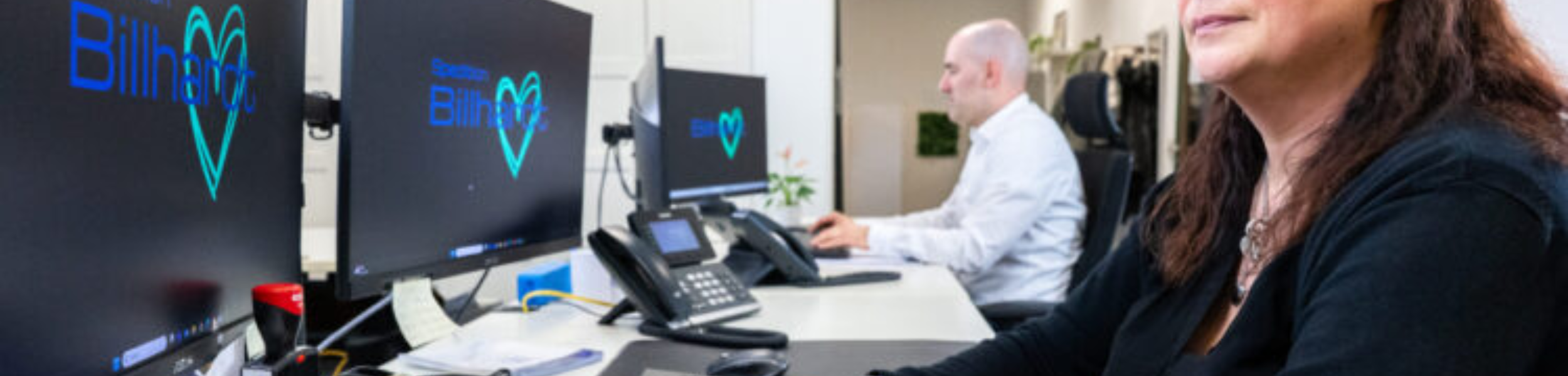 Zwei Personen arbeiten an Computern in einem Büro mit Bildschirmen, die ein Herzsymbol und den Schriftzug "Blitz" zeigen.